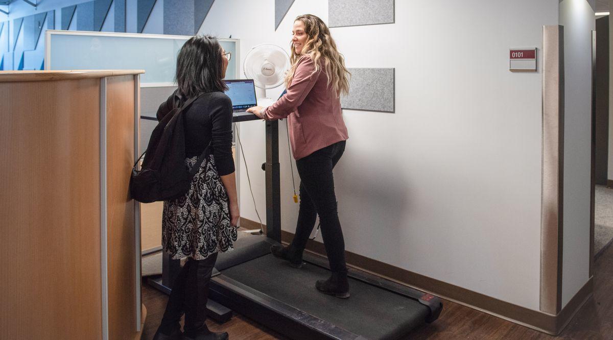 A woman and a man stand while having a discussion in front of an elevated work desk