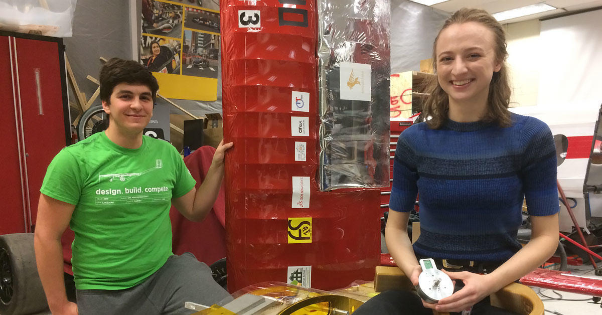 Matt Sehri est agenouillé près d’un modèle d’aéronef, une main posée sur son aile. Sarah Horton, souriante, est assise sur un tabouret et tient à la main un moteur et un membre robotisé recourbé. 