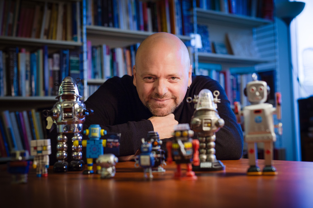 a man is sitting at a desk. His arms are stacked and rested on the desk and his chin is rested on top of his hands. There are small robots in the foreground, on the desk.