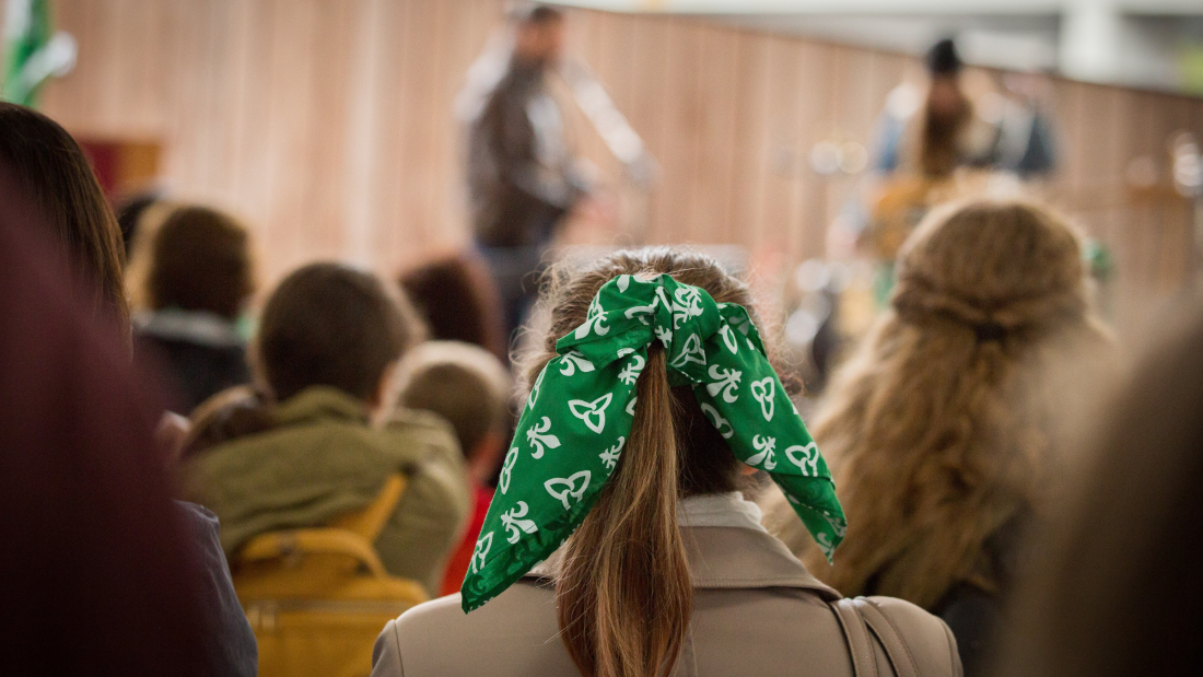 The photo depicts a Canadian conference with a young woman wearing a green headband symbolizing the Francophonie, surrounded by attentive spectators.