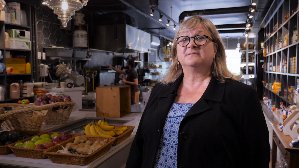 A woman stands in the foreground of a food market. There are trays of fruits and shelves of non-perishable items behind her.