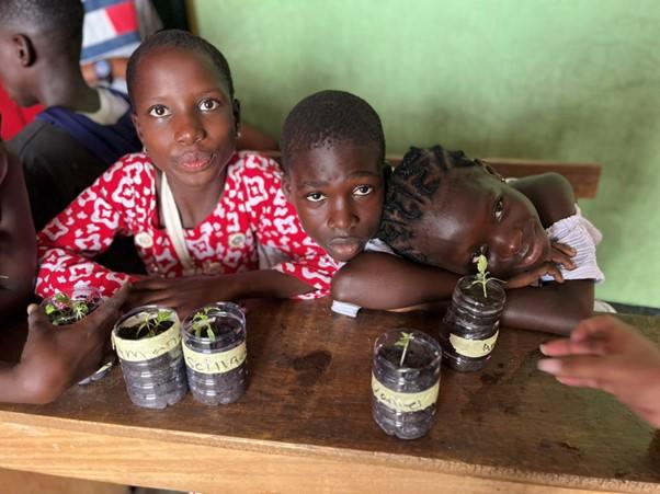 Three middle school kids sitting at a table in a classroom. 4 bottles on the table each containing a small plant