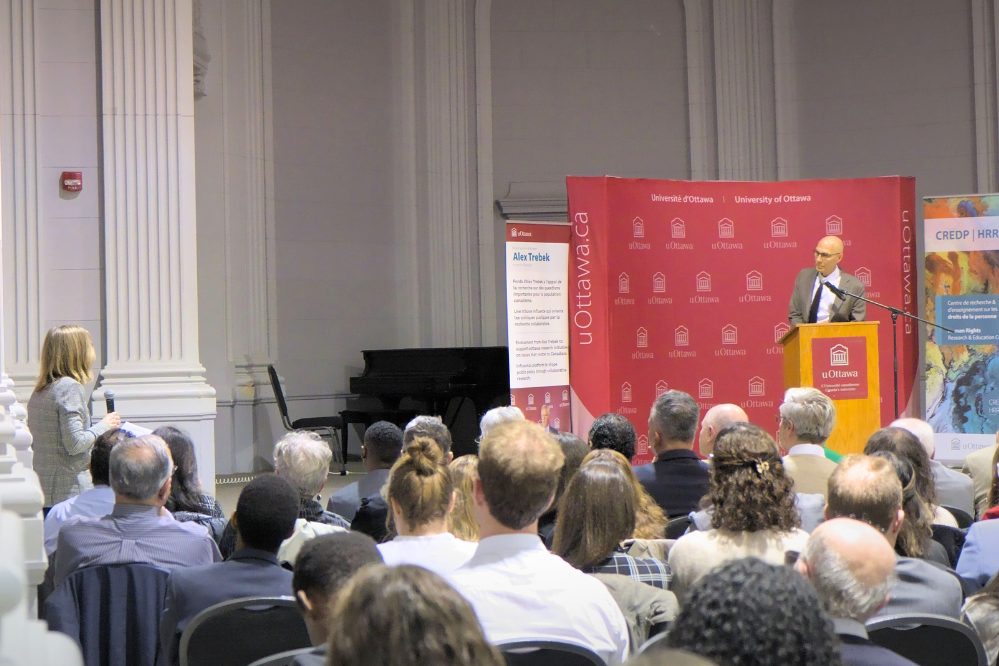 A wide view of a room where many people are seated, facing a man at a podium in the distance. There is a woman standing in the audience with a microphone. She and the speaker are facing each other.