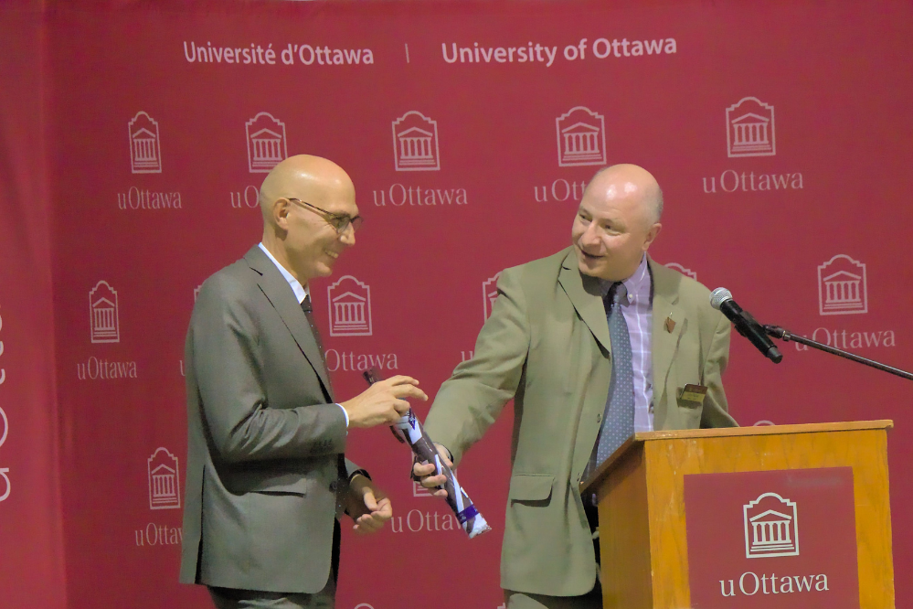 two  men stand at a podium. One is extending his hand to the other, offering a gift