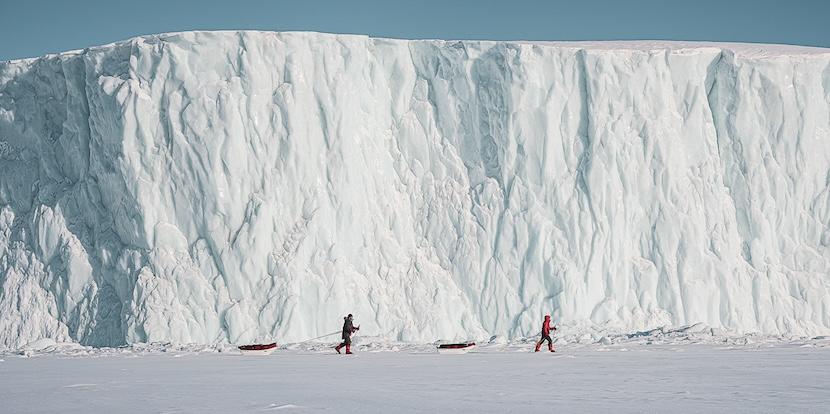 Two explorers pulling their sleds in front of an icy cliff face