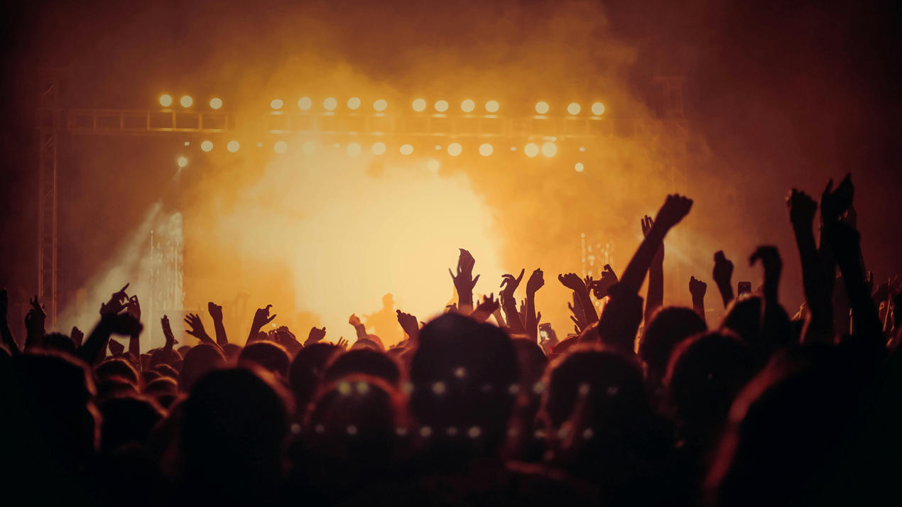 A crowd with hands raised in silhouette in front of a brightly lit stage.