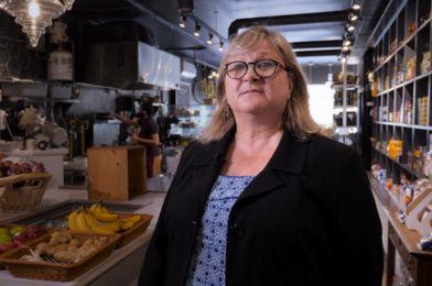 A woman stands in the foreground of a food market. There are trays of fruits and shelves of non-perishable items behind her.