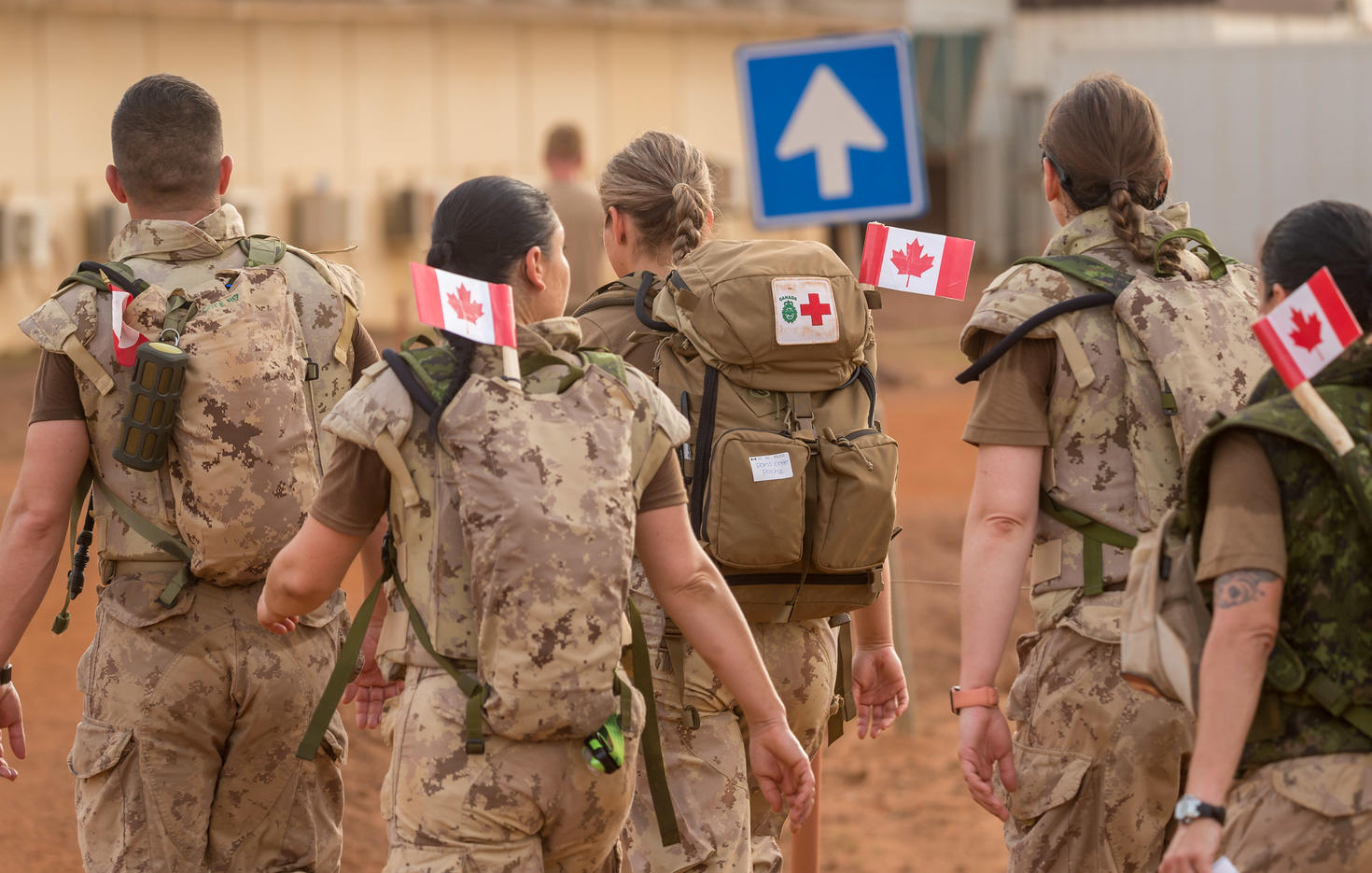 Military personnel in camouflage uniforms walk away, each carrying backpacks with Canadian flags attached.