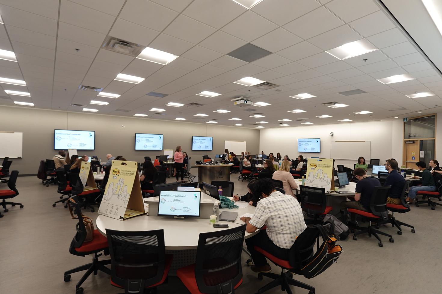 A modern classroom with multiple individuals seated at round tables, each working on laptops or monitors.