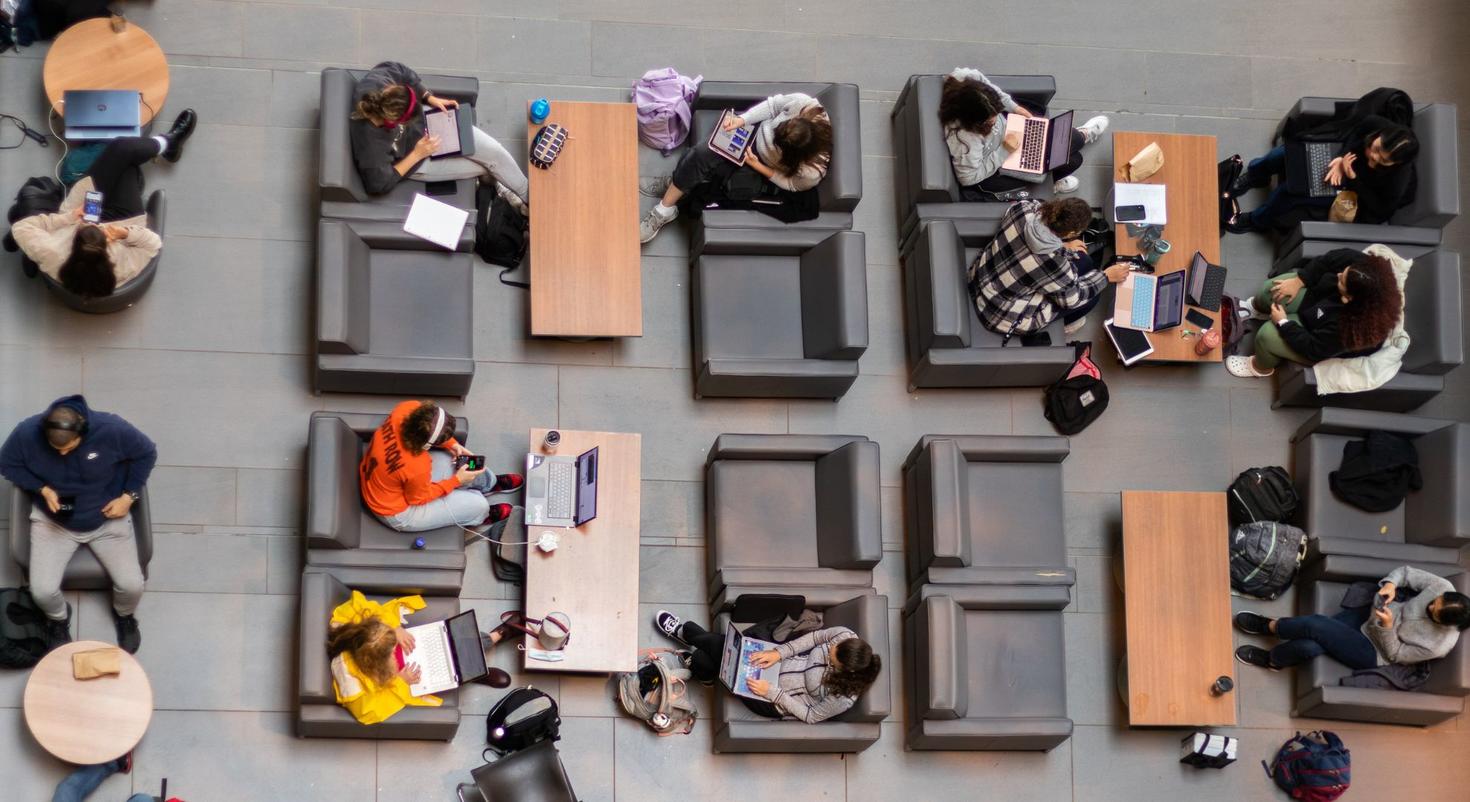  A group of students sitting in a common area, using laptops and tablets while seated in chairs around tables. The scene is viewed from above.
