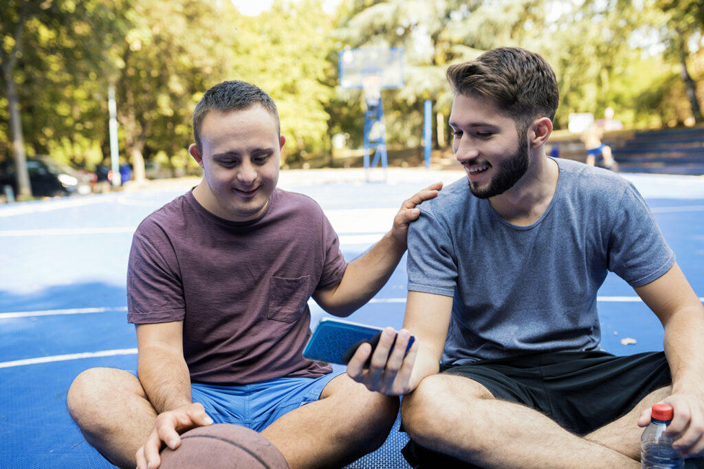 Two people sitting on a blue sports court, one holding a basketball and the other a phone, engaged in conversation.