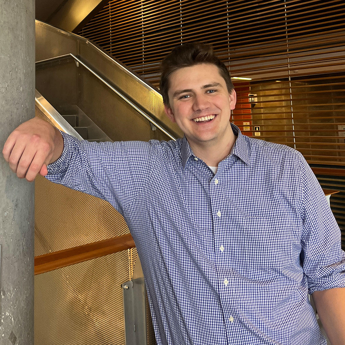 Joe Workentin smiles while leaning against a concrete pillar in a modern building with wooden accents and a staircase in the background.