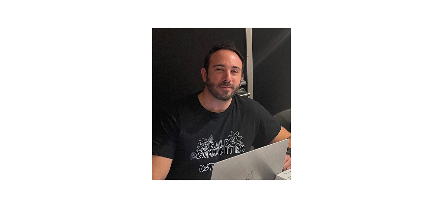 A person with short dark hair and a beard sits at a table with a laptop, wearing a black t-shirt with "Build Communities" printed on it.