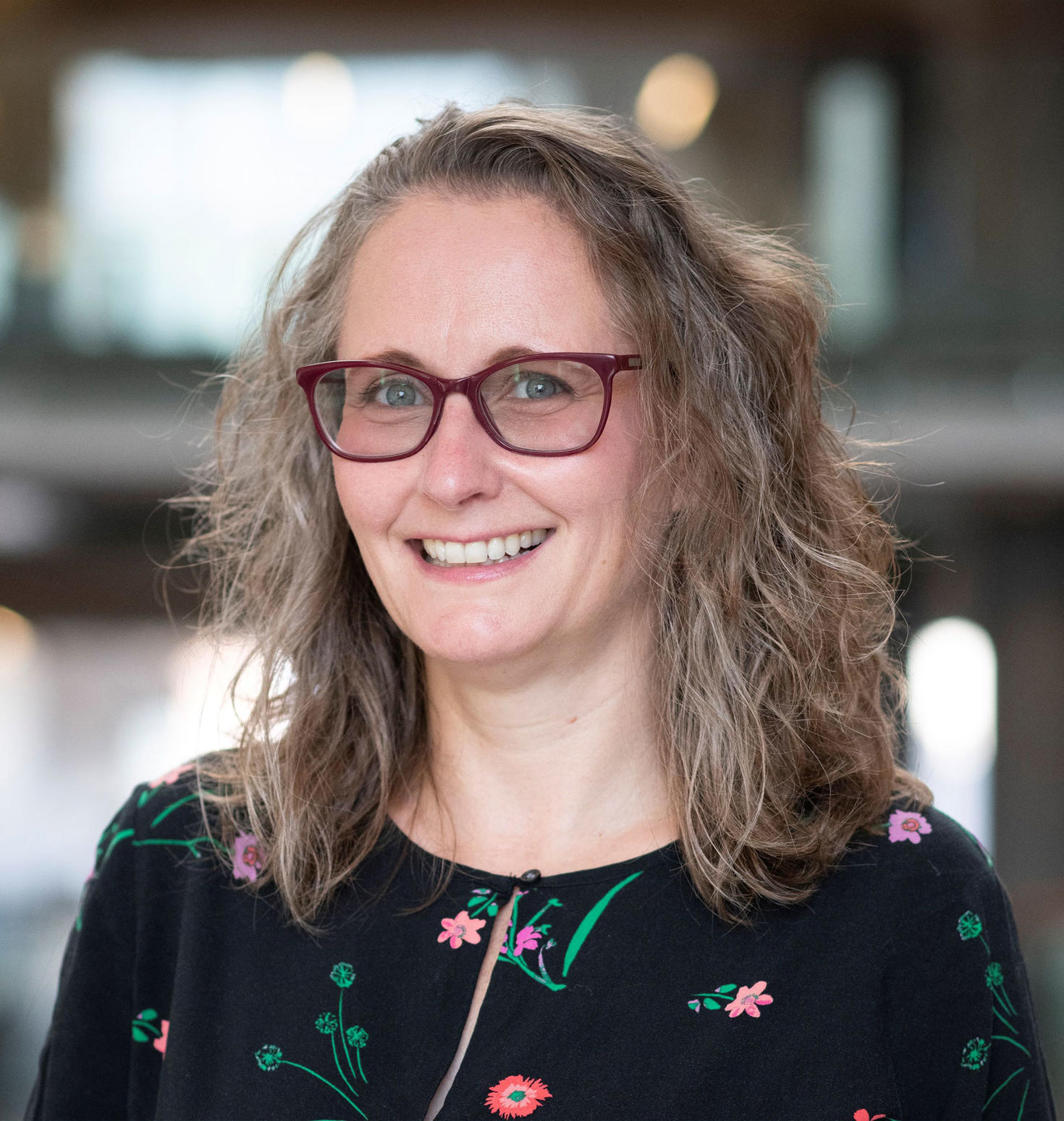 Portrait of Professor Shanna Kousaie smiling, wearing glasses and a floral-patterned top, with a blurred indoor background.
