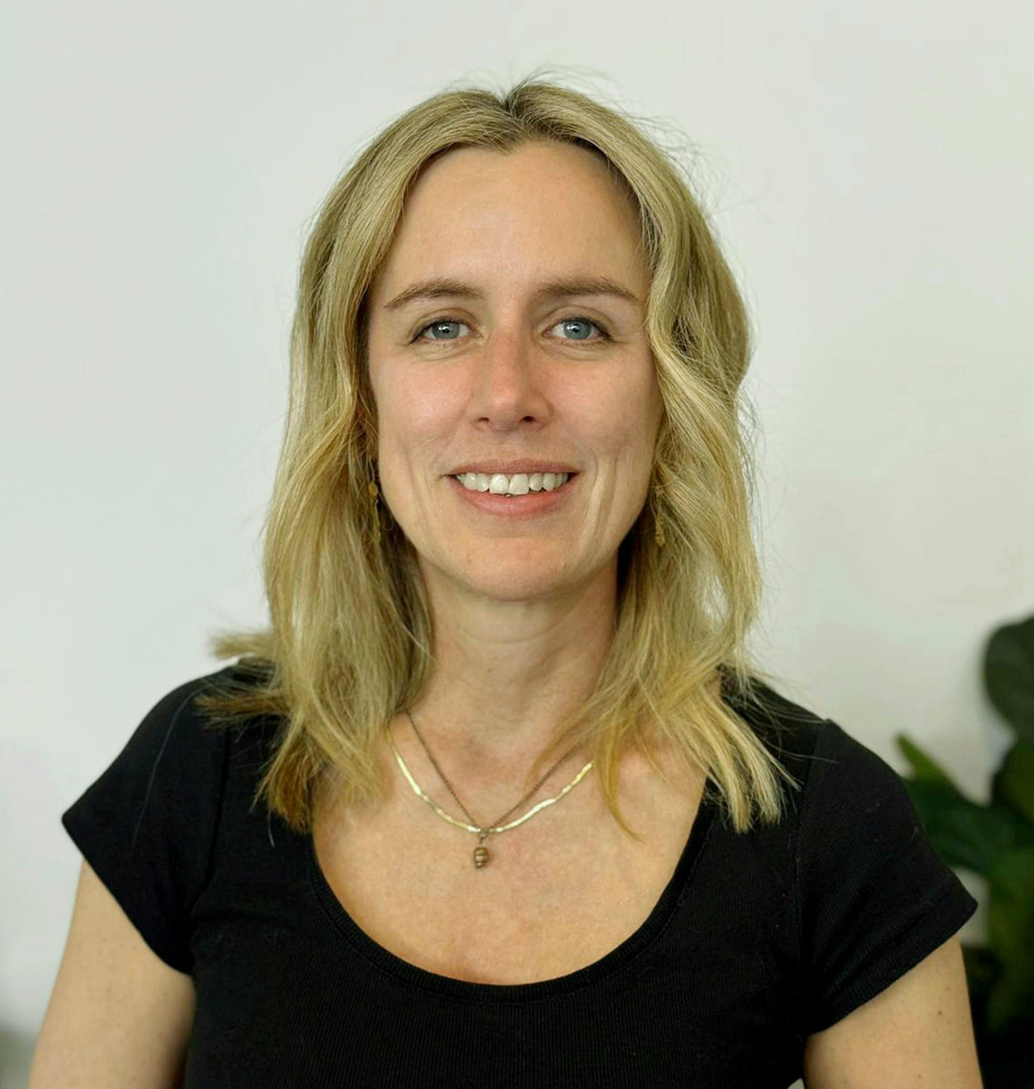 Portrait of Professor Vanessa Taler smiling, wearing a black top, with a plain background and a plant partially visible.