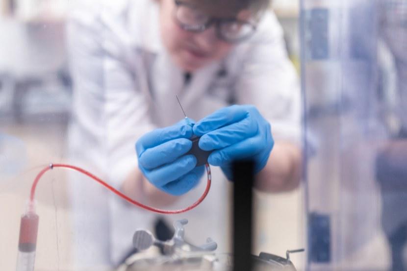 A researcher wearing a white lab coat, safety glasses, and blue gloves carefully handles a syringe and tubing in a laboratory setting.