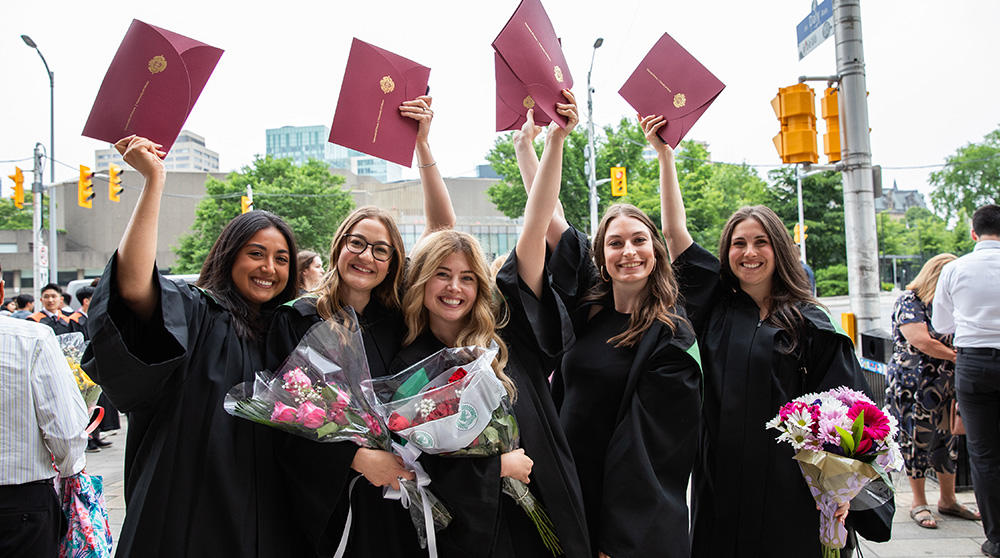 Five women in graduation gowns hold up their diplomas.