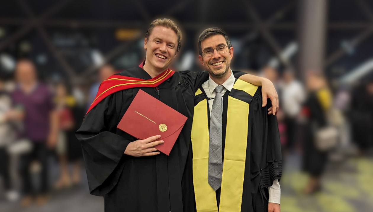 : Adam Cook in a black gown with a red and gold hood, holding a diploma. To his left stands Professor Stephen Newman, in a black gown with a yellow stole. They’re surrounded by others celebrating graduation. 
