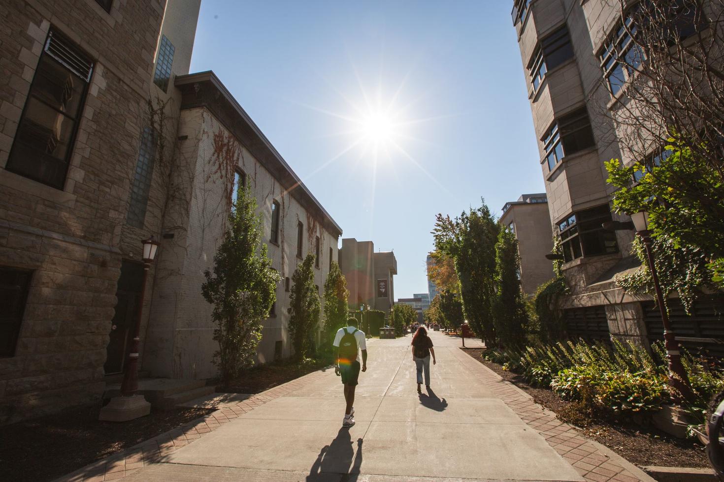 Two students walk past campus buildings and lush green trees in the bright sunshine.
