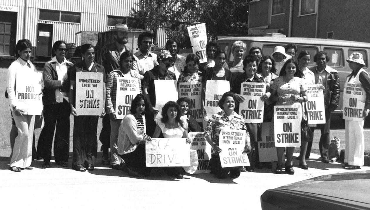Workers on strike displaying “Upholsterers International Union-Local 1” and “On Strike” signs