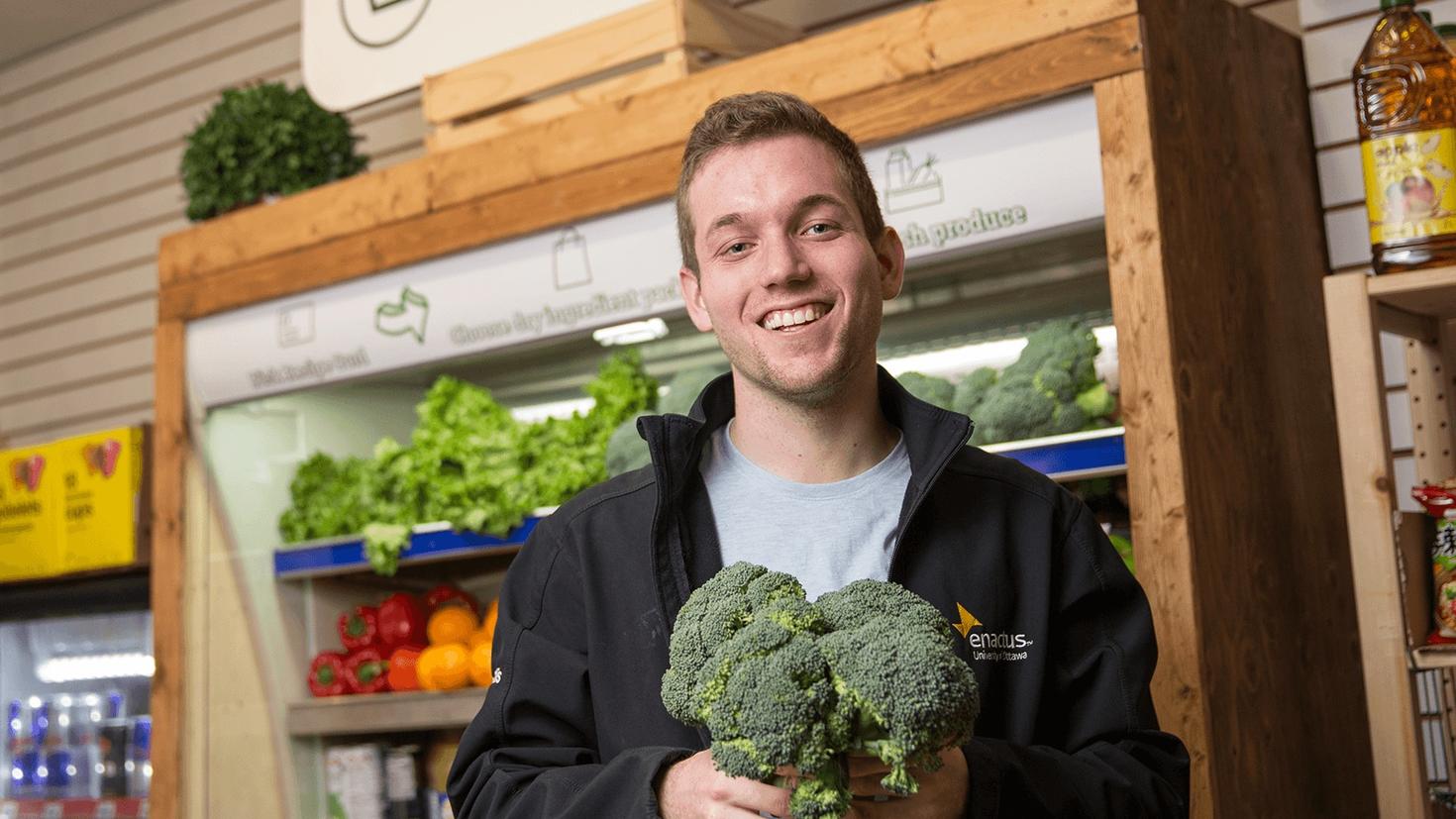 Corey Ellis, smiling and holding some broccoli, stands in front of a refrigerated unit full of produce.