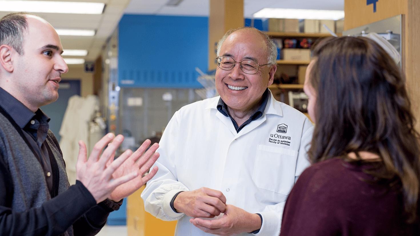 Dressed in a white lab coat and with a glowing smile, Zemin Yao chats with two people.
