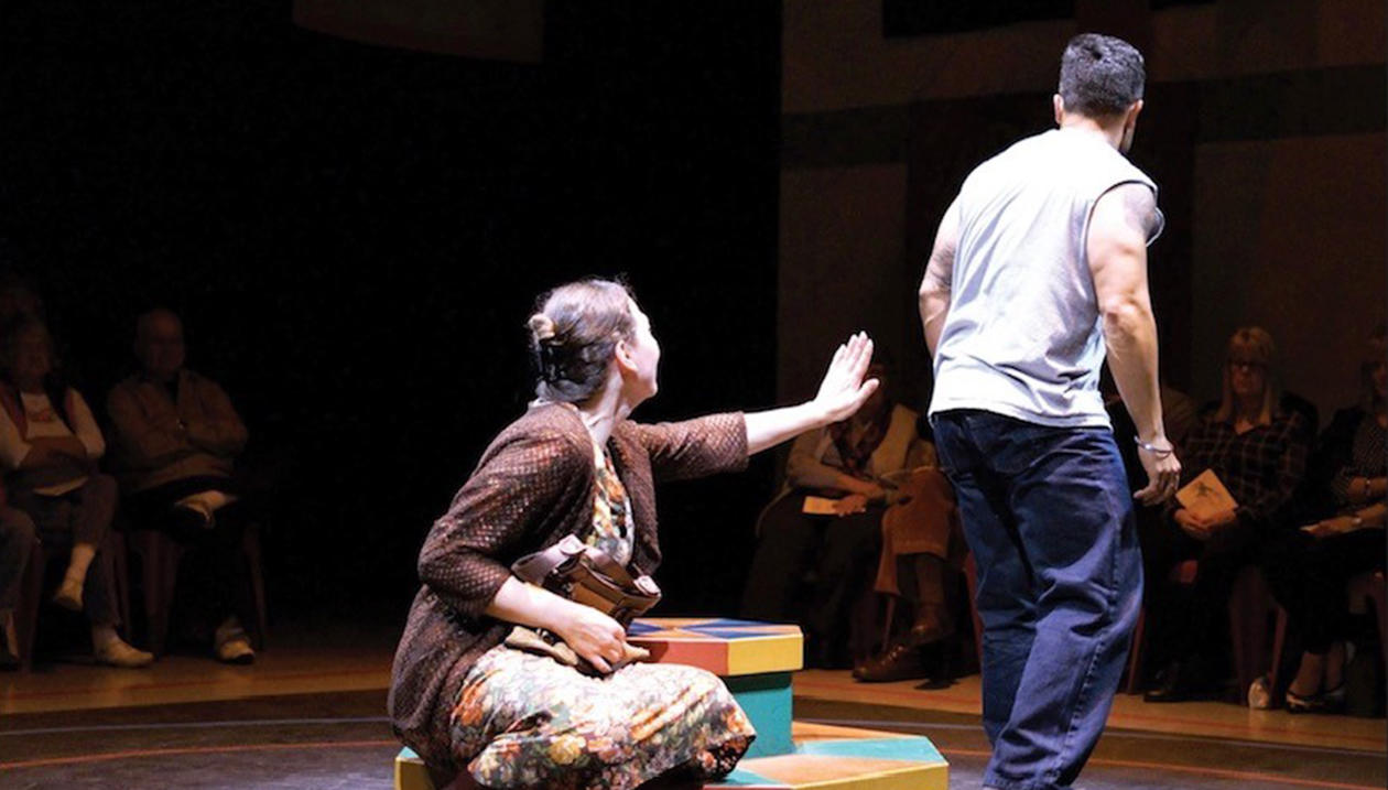 A seated woman reaches toward a departing man on stage as the audience watches.