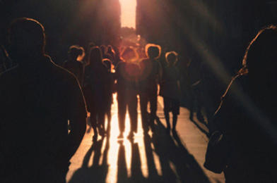 Silhouetted crowd walks down a sunlit street casting long shadows.