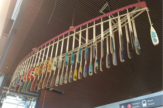 A canoe-shaped frame hanging from a ceiling in a train station, with colourful canoe paddles hanging from the frame