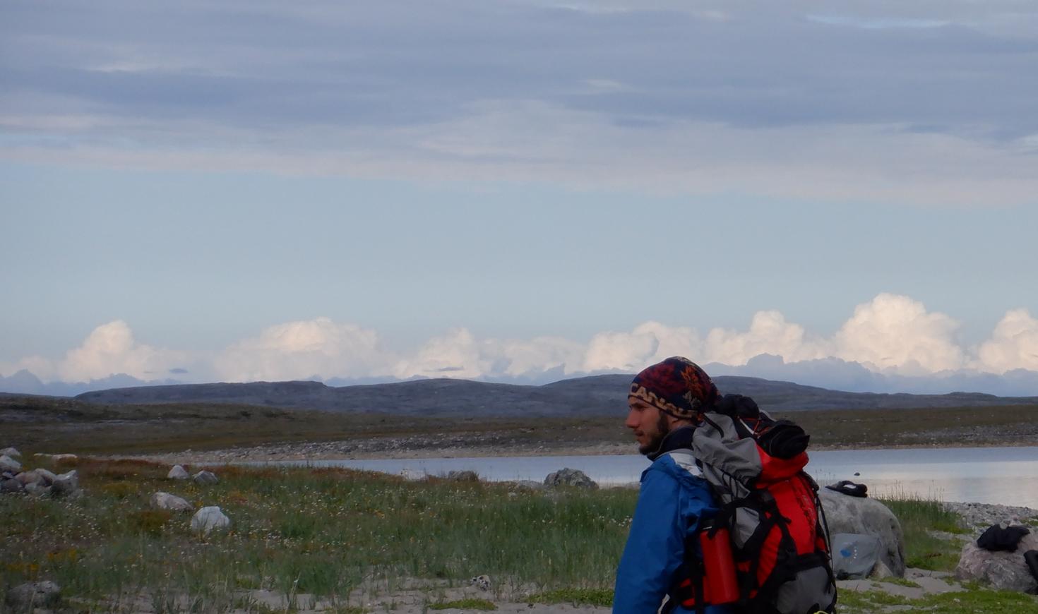 PhD student Victor Garcia in the field stands with a large hiking backpack beside a remote Arctic lake near Inukjuak, northern Quebec, with Hudson Bay in the background. 