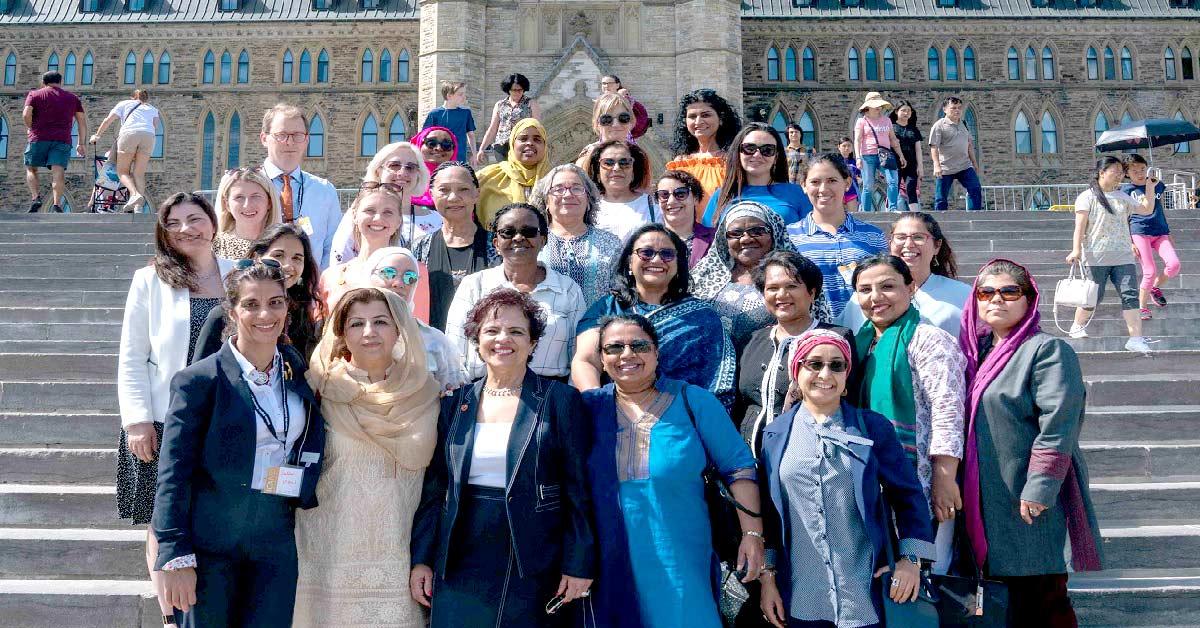 Two dozen people, mostly women, stand on the steps in front of Centre Block.