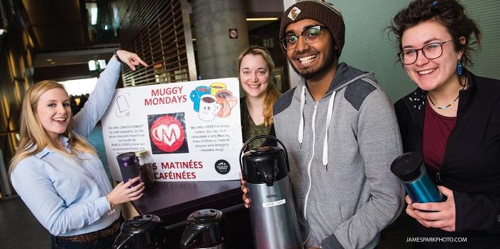 Three women and a man hold reusable coffee mugs, one woman is pointing at a Muggy Mondays sign.