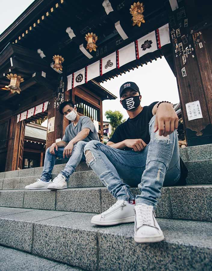 Two men wearing surgical-type face masks sit on steps in front of an ornate wooden structure