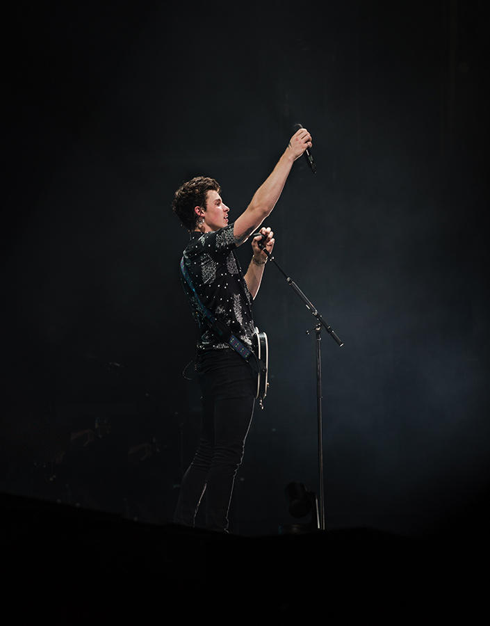 Shawn Mendes with a guitar strapped on and holding up a microphone.