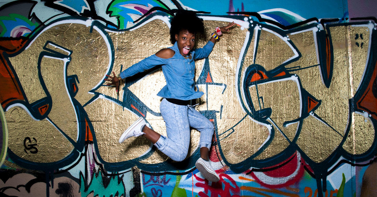 Pearline Barrett-Fraser is jumping and dancing in front of a wall covered with colourful grafifitis