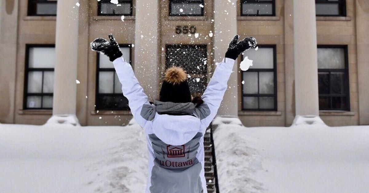 Un individu portant un manteau avec le logo de l'Université d'Ottawa jette de la neige en l'air, celui-ci a été photographié par derrière.