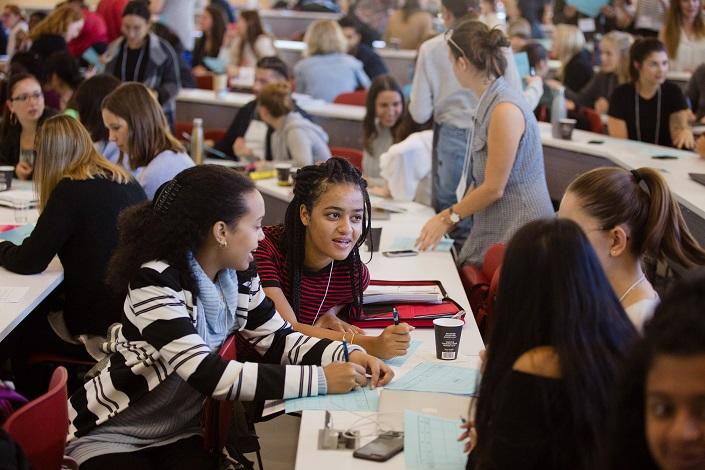 Four yo0ung women engage in conversation in a classroom. Other students chat in groups behind them.