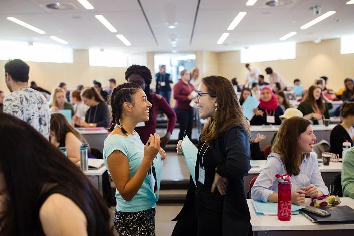 Two female students stand together, smiling and talking, at the front of a classroom. Other students behind them also chat in groups.